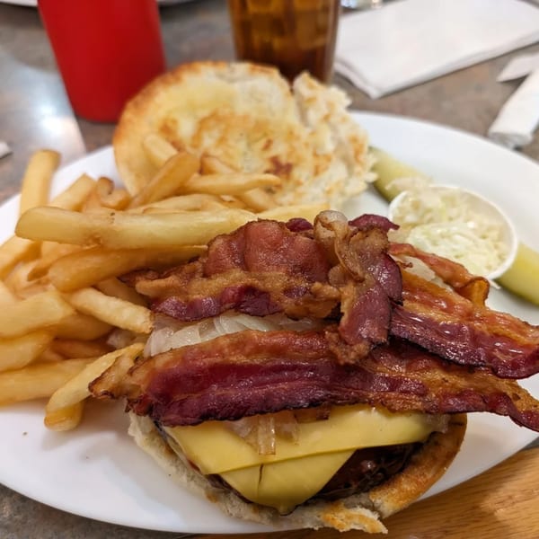 A bacon cheeseburger with fries, a pickle spear, and coleslaw on a white plate at Old Glory.