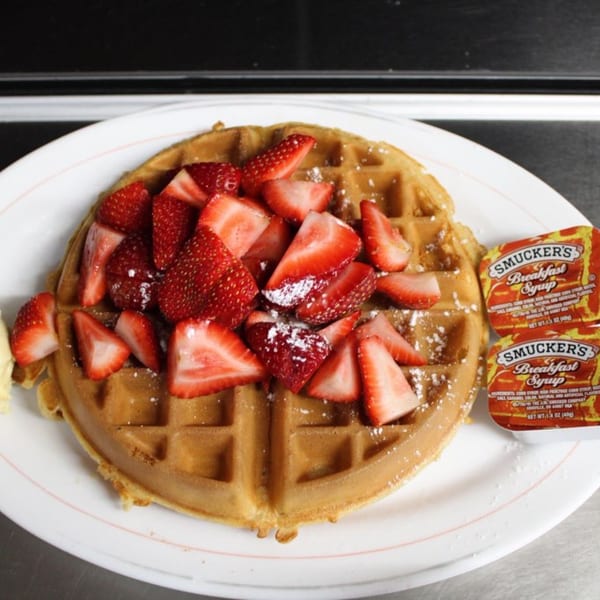 A golden waffle topped with sliced strawberries and powdered sugar, with two syrup packets.