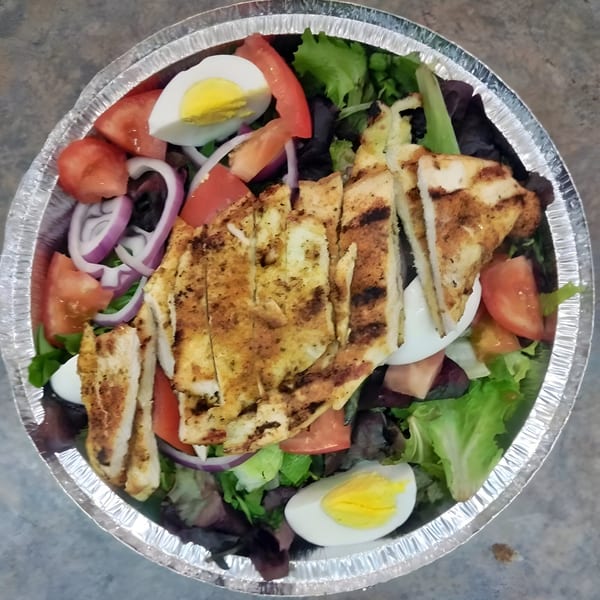 Grilled chicken salad with lettuce, tomatoes, red onions, and hard-boiled eggs in a foil container.