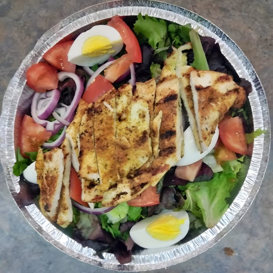 Grilled chicken salad with lettuce, tomatoes, red onions, and hard-boiled eggs in a foil container.