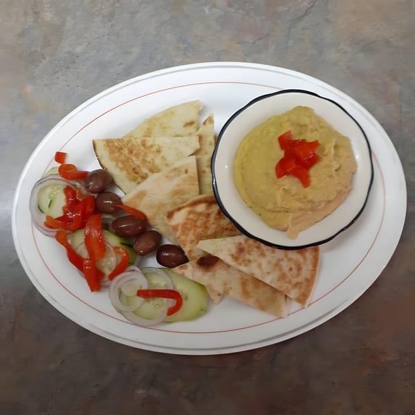Hummus with pita bread, olives, sliced cucumbers, onions, and red peppers on a white plate.