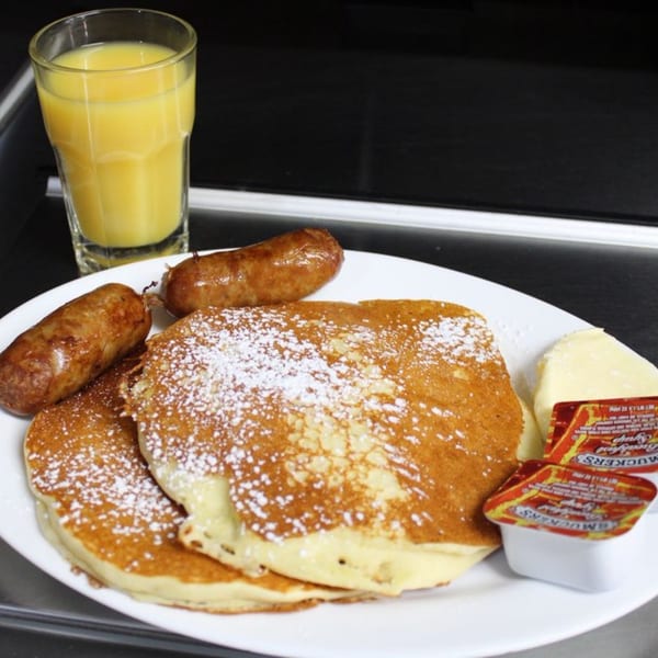 Pancakes with powdered sugar, two sausages, butter, syrup, and a glass of orange juice.